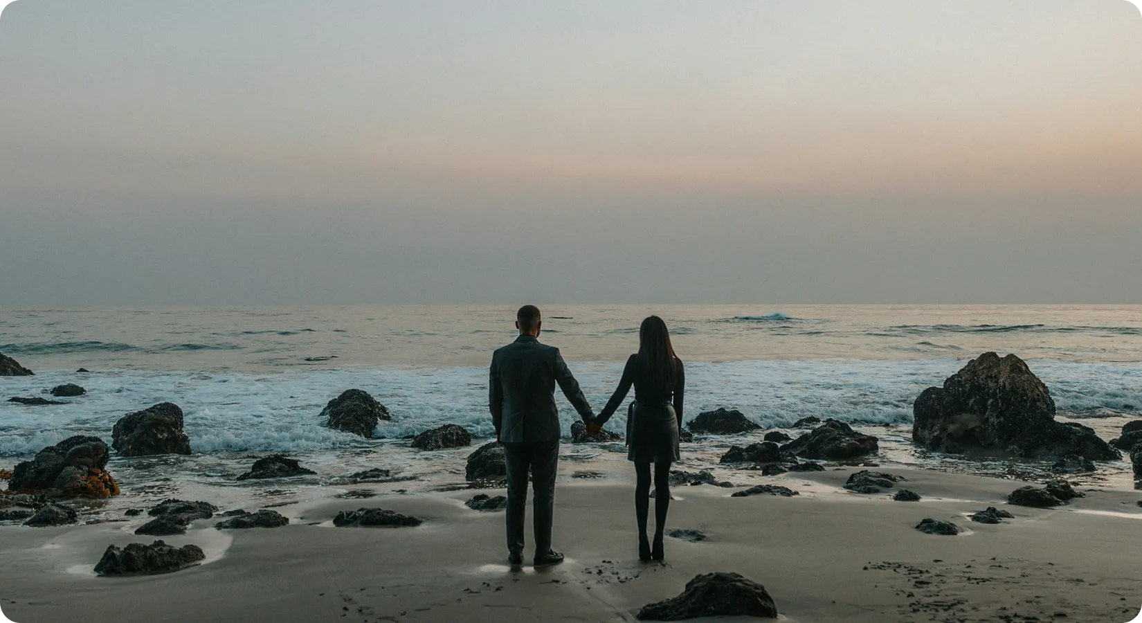 A couple holding hands on a rocky beach, looking out at the sea at dusk