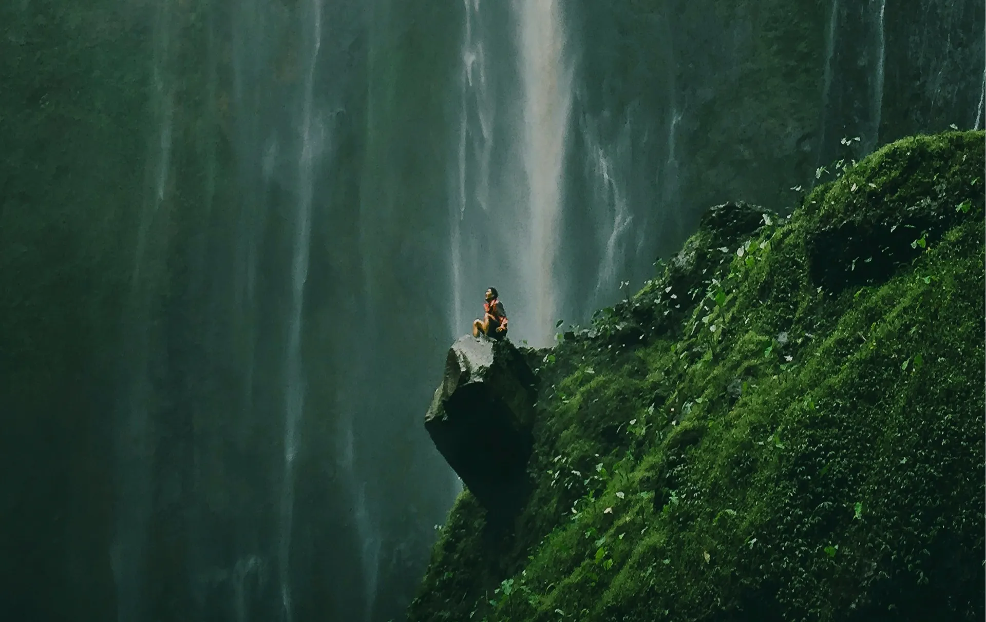 A person sitting on a mossy cliff edge beside a tall waterfall