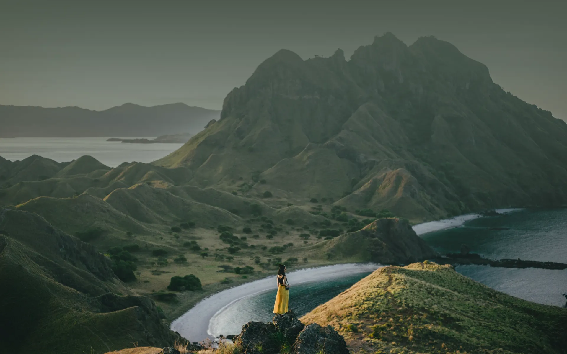 A woman in a yellow dress standing on a ridge above green hills and a winding bay