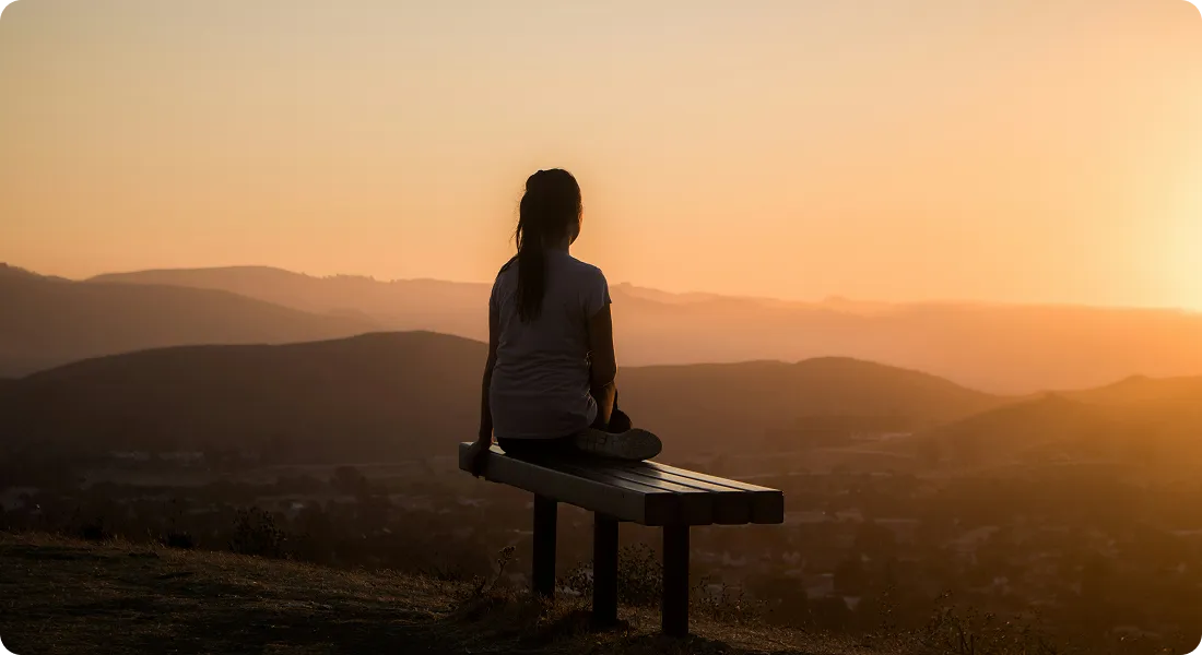 A woman sitting alone on a bench, looking out over the hills at sunset