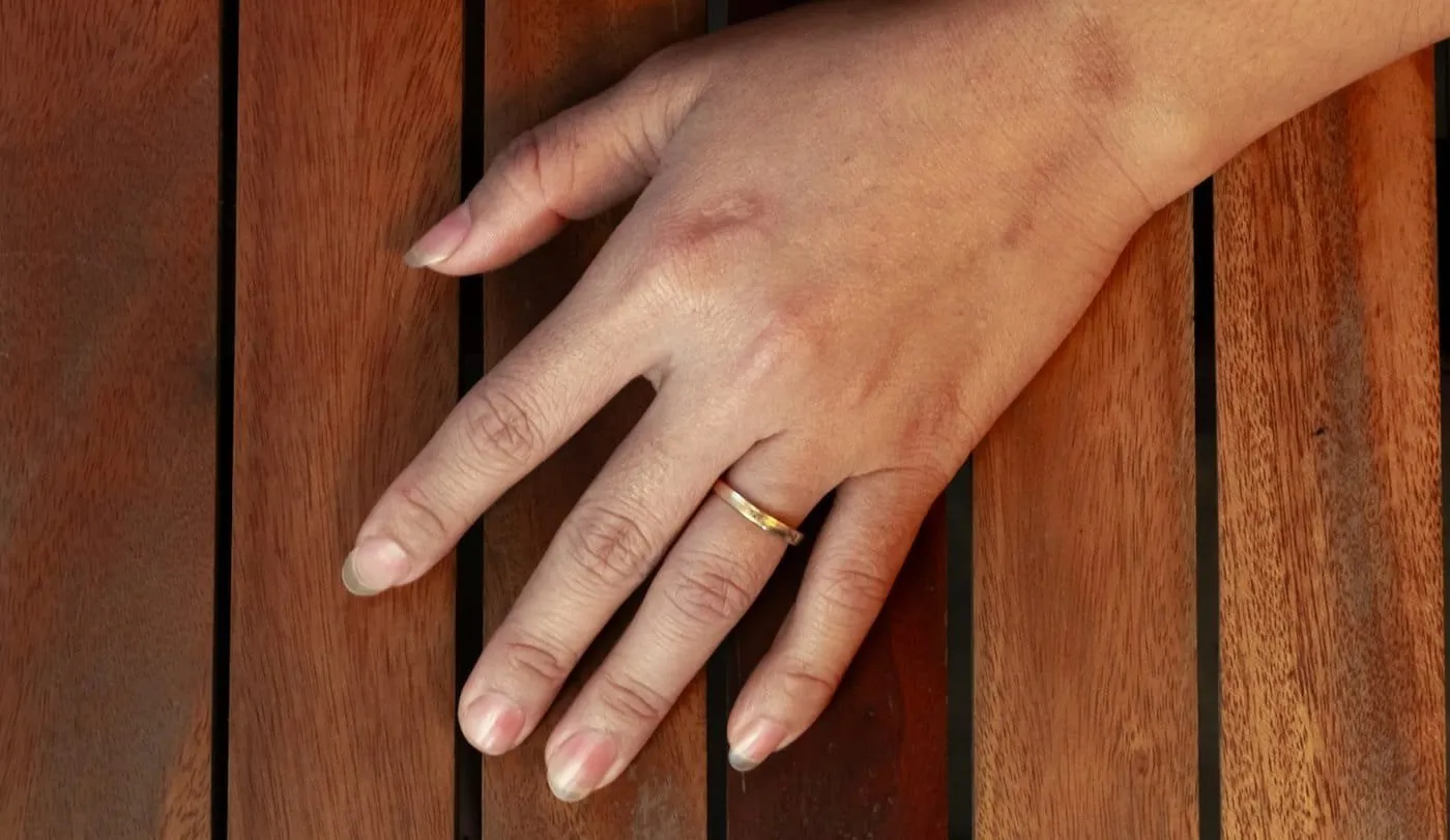 A hand wearing a gold wedding ring resting on warm wooden slats in afternoon light