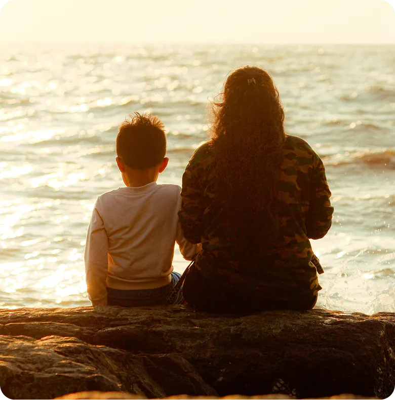 A mother and her young son sitting together on the rocks, looking out at the sea at sunset